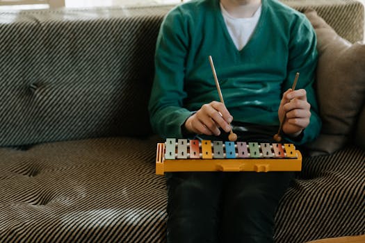 A child in a green sweater plays a colorful xylophone while sitting on a sofa indoors.